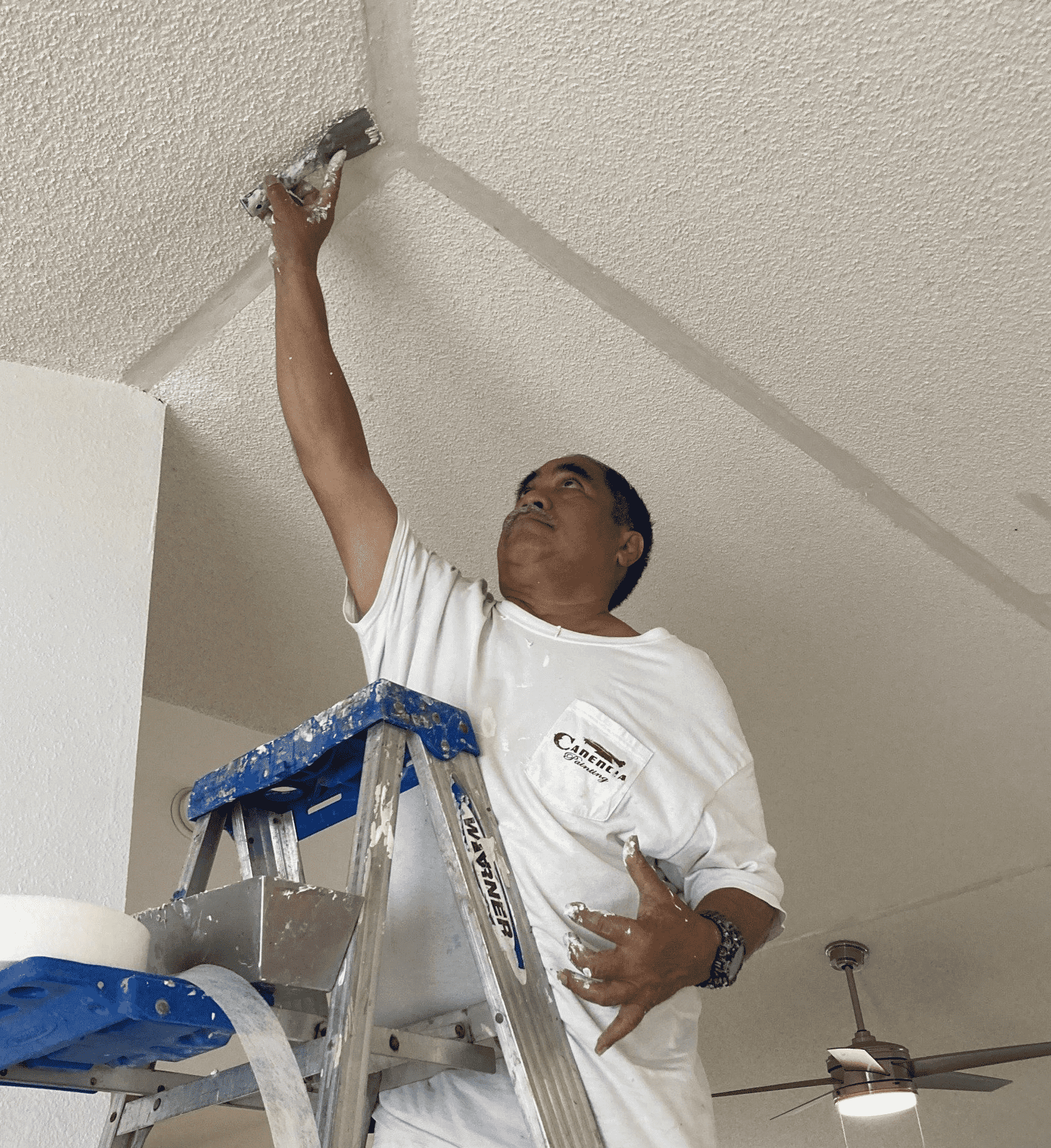 Bryan Canencia painting a ceiling in white painter's attire on a ladder inside an Oahu home